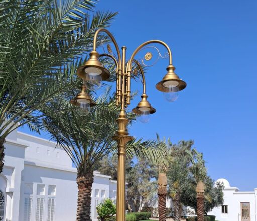 Rikt dekorerad gatlykta i guldfärgad nyans med fyra armaturer 04/Aries/A, mot en bakgrund av palmer och vita byggnader en solig dag